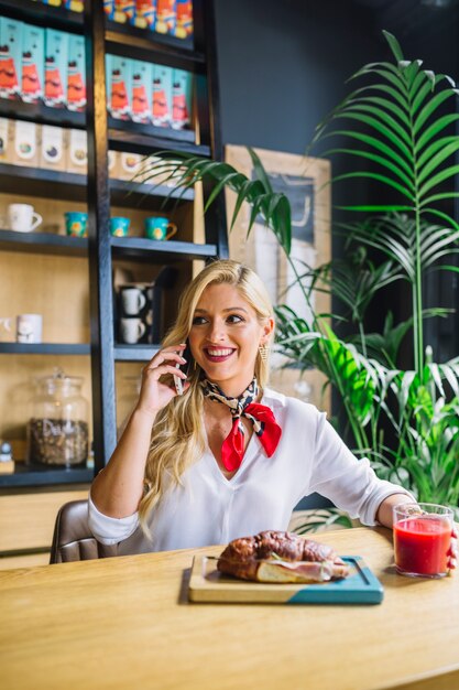 Giovane donna bionda che parla sul telefono cellulare che mangia vetro di succo e di pane al forno