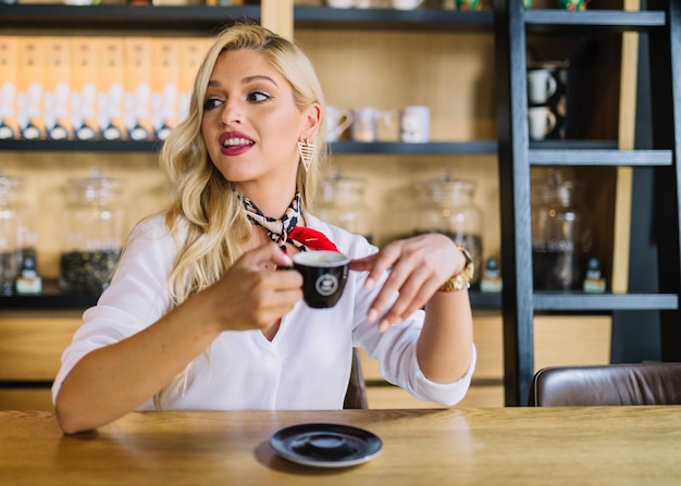 Giovane donna bionda che si siede nel caffè che tiene distogliere lo sguardo della tazza di caffè