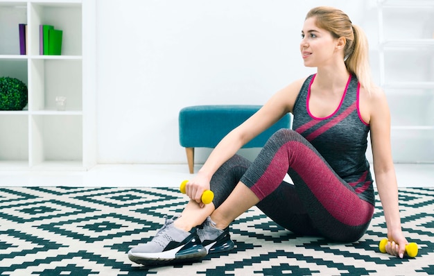 Giovane donna bionda che si siede sul tappeto a casa tenendo dumbbells gialli in mano