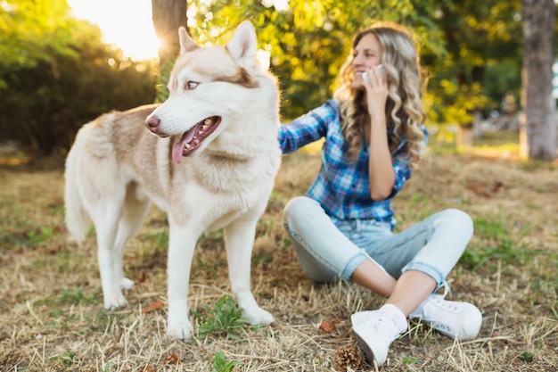 Giovane donna bionda felice abbastanza sorridente che gioca con la razza del cane husky nel parco il giorno di estate pieno di sole