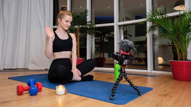 Giovane donna bionda in abiti sportivi su una stuoia di yoga salutando la videocamera di registrazione davanti a lei