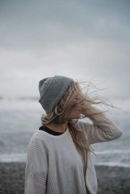 Giovane donna bionda in depressione con un cappello che cammina sulla spiaggia in un tempo ventoso