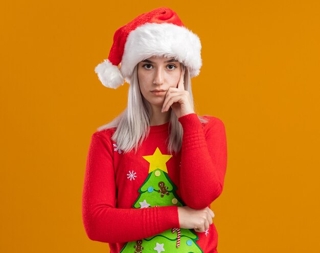 giovane donna bionda in maglione di Natale e cappello da Babbo Natale guardando la fotocamera con la faccia seria in piedi su sfondo arancione