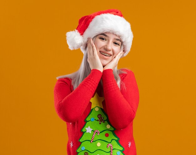 Giovane donna bionda in maglione di Natale e cappello della Santa che guarda l'obbiettivo sorridente felice e positivo allegramente in piedi su sfondo arancione