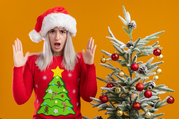 Giovane donna bionda in maglione di natale e cappello di babbo natale con la faccia arrabbiata che alza le mani in piedi accanto a un albero di natale sopra il muro arancione