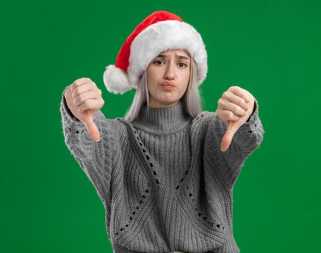 Giovane donna bionda in maglione invernale e cappello da Babbo Natale che guarda l'obbiettivo di essere dispiaciuto che mostra i pollici verso il basso in piedi su sfondo verde