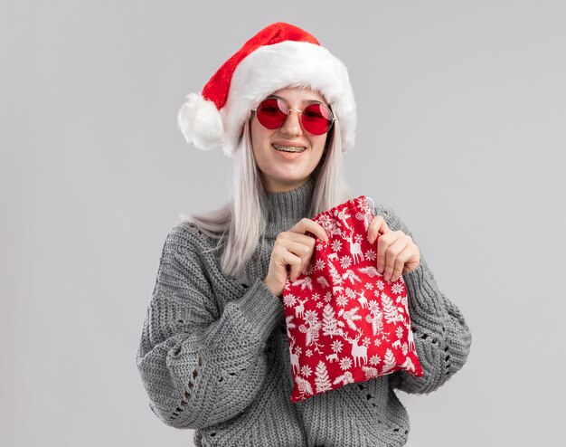Giovane donna bionda in maglione invernale e cappello da Babbo Natale che tiene in mano una borsa rossa con regali di Natale con un sorriso sul viso in piedi su un muro bianco white