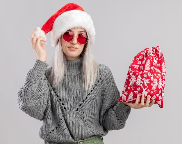 Giovane donna bionda in maglione invernale e cappello da Babbo Natale che tiene la borsa rossa di Babbo Natale con regali di Natale felice e gioioso che guarda da parte in piedi sul muro bianco