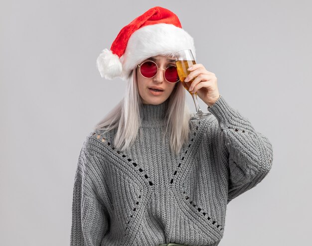 Giovane donna bionda in maglione invernale e cappello da Babbo Natale che tiene un bicchiere di champagne in cerca di mal di testa con mal di testa