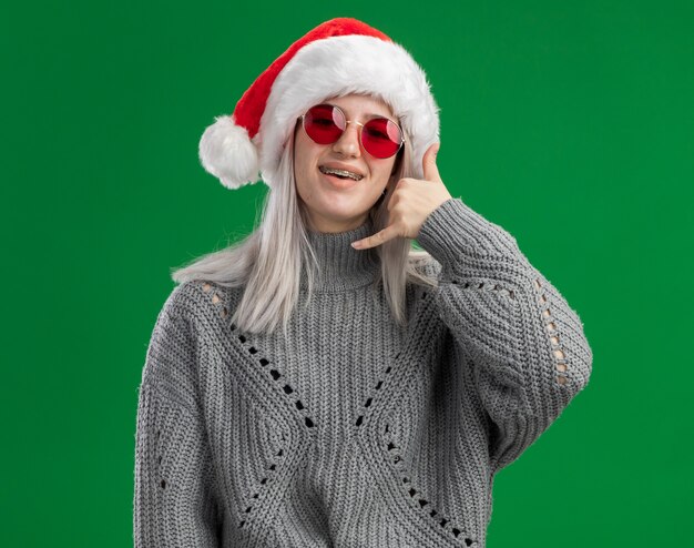 Giovane donna bionda in maglione invernale e cappello da Babbo Natale con gli occhiali rossi che guarda l'obbiettivo felice e allegro che mi chiama gesto in piedi su sfondo verde
