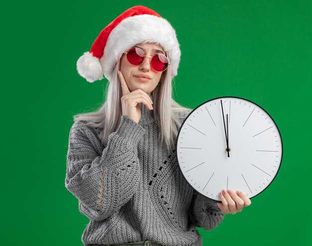Giovane donna bionda in maglione invernale e cappello da Babbo Natale con gli occhiali rossi che tengono l'orologio da parete con un'espressione pensierosa che pensa in piedi sul muro verde