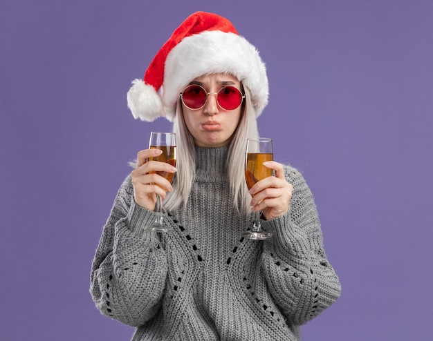 Giovane donna bionda in maglione invernale e cappello da Babbo Natale con in mano due bicchieri di champagne con un'espressione triste che arriccia le labbra in piedi sul muro viola