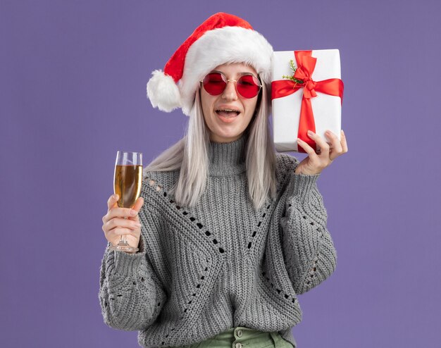 Giovane donna bionda in maglione invernale e cappello da Babbo Natale con in mano un regalo e un bicchiere di champagne felice ed eccitata in piedi sul muro viola