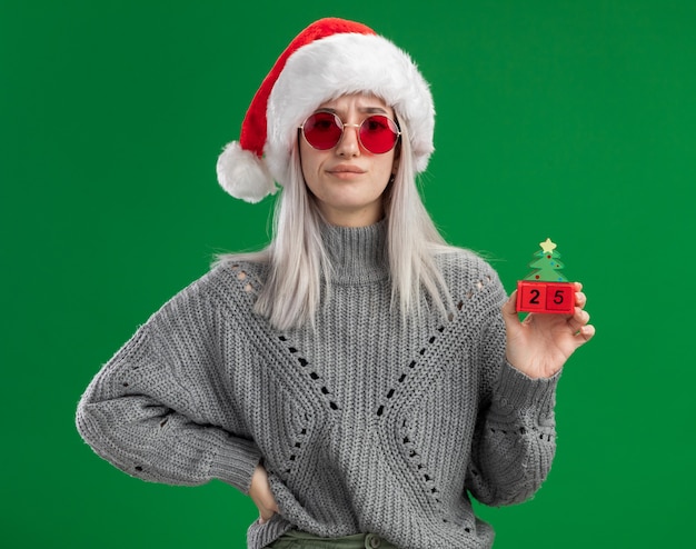 Giovane donna bionda in maglione invernale e santa hat indossando occhiali rossi tenendo i cubi giocattolo con la data di natale guardando la telecamera essendo scontento in piedi su sfondo verde