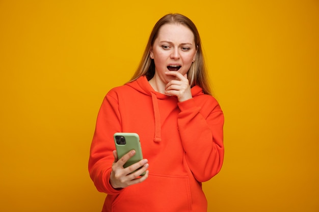 Giovane donna bionda interessata che tiene e che esamina il telefono cellulare che tiene la mano sul mento