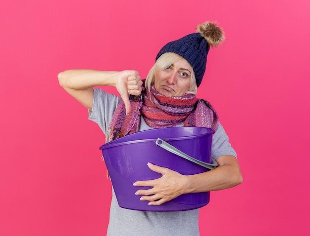 Giovane donna bionda malata slava dispiaciuta che indossa cappello invernale e sciarpa che tiene secchio di plastica e pollice in giù isolato sulla parete rosa con lo spazio della copia