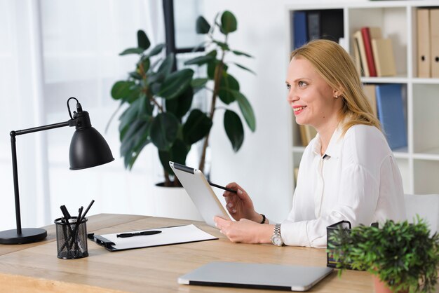 Giovane donna bionda sorridente che si siede nel luogo di lavoro facendo uso della compressa digitale