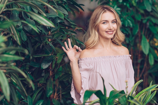 Giovane donna bionda sorridente che sta davanti alla pianta verde che mostra segno giusto