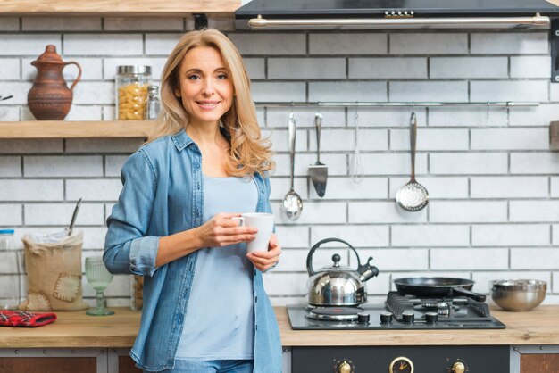 Giovane donna bionda sorridente che sta vicino alla stufa di gas che tiene la tazza di caffè macchiato