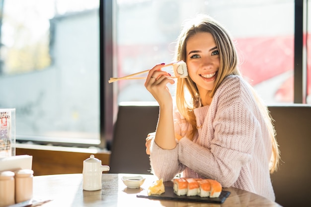 Giovane donna bionda sorridente piena di sole in maglione bianco che mangia sushi per pranzo in un piccolo caffe