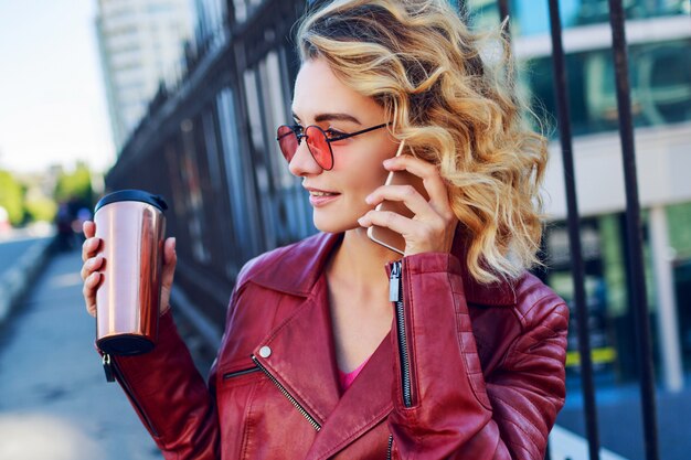 Giovane donna bionda vaga che cammina nella città e che per mezzo dello Smart Phone. Dettagli da vicino. Ragazza moderna alla moda con caffè. Peli ventosi.