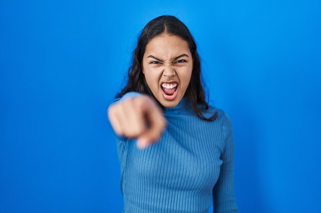 Giovane donna brasiliana in piedi su sfondo blu isolato che punta scontento e frustrato verso la telecamera, arrabbiata e furiosa con te