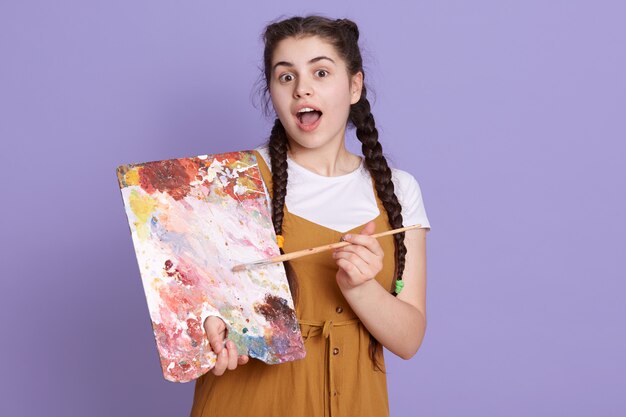 Giovane donna bruna artista con le trecce che tiene il pennello e la tavolozza del pittore sulla parete lilla, in posa con la faccia a sorpresa, in piedi con la bocca aperta.