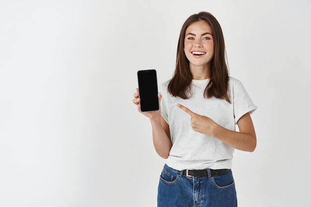 Giovane donna bruna che mostra lo schermo vuoto dello smartphone e punta il dito verso il telefono cellulare sorridendo soddisfatto alla telecamera in piedi su sfondo bianco