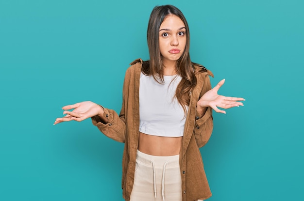 Giovane donna castana che indossa abiti casual all'oscuro e espressione confusa con le braccia e le mani sollevate concetto di dubbio