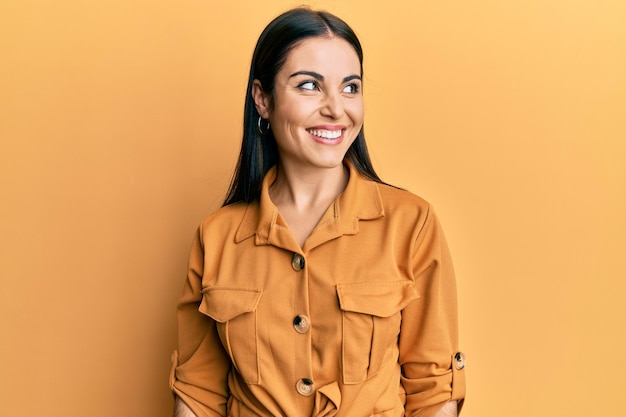 Giovane donna castana che indossa abiti casual che distoglie lo sguardo a lato con il sorriso sul viso, espressione naturale. ridendo fiducioso.