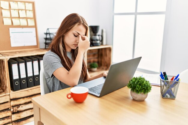 Giovane donna castana che lavora in ufficio con il laptop stanco sfregamento naso e occhi sensazione di affaticamento e mal di testa stress e concetto di frustrazione