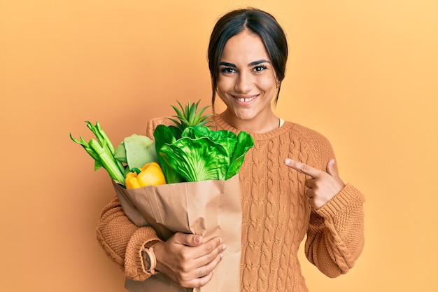 Giovane donna castana che tiene il sacchetto di carta con pane e generi alimentari sorridendo felice indicando con mano e dito