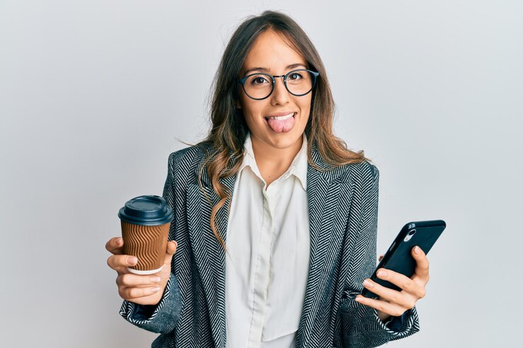 Giovane donna castana che usa lo smartphone e beve una tazza di caffè con la lingua fuori felice con un'espressione divertente.