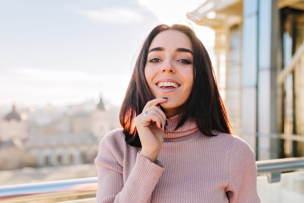 Giovane donna castana gioiosa del ritratto del primo piano che si diverte sulla terrazza nella mattina soleggiata sulla vista della grande città. Stato d'animo allegro, eccitato, momento di relax, sorridente.