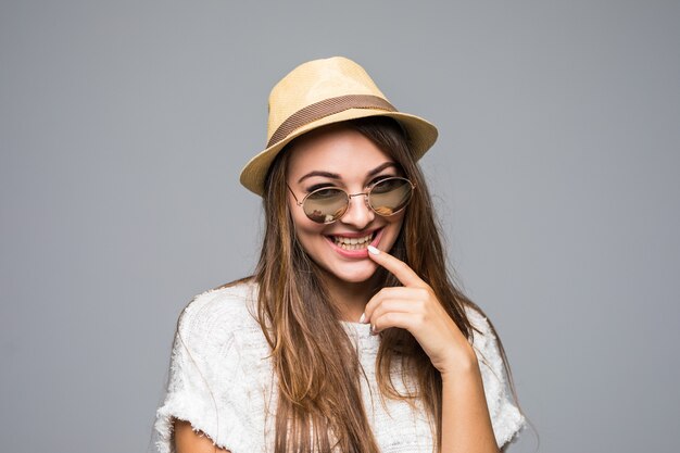 Giovane donna castana in cappello e occhiali da sole che sorride su gray