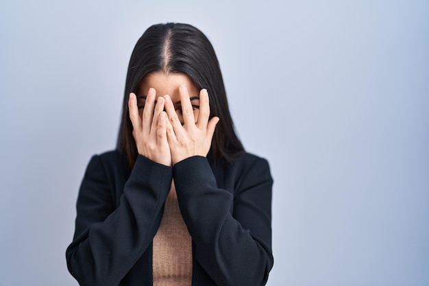 Giovane donna castana in piedi su sfondo blu con espressione triste che copre il viso con le mani mentre piange il concetto di depressione