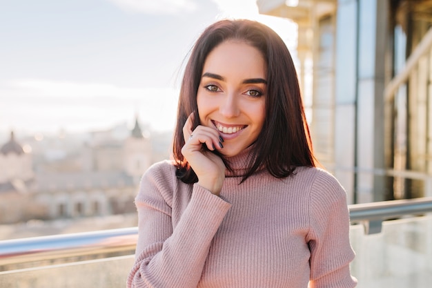 Giovane donna castana sorridente gioiosa del ritratto nella mattina soleggiata agghiacciante sulla terrazza. Sorridere, esprimere emozioni positive del viso, successo, divertimento.