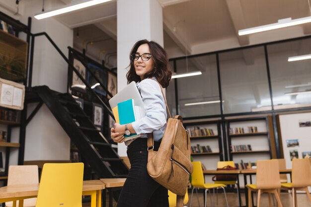Giovane donna castana sorridente in vetri neri che cammina con roba di lavoro e laptop in biblioteca. Studente intelligente, vita universitaria, sorridente