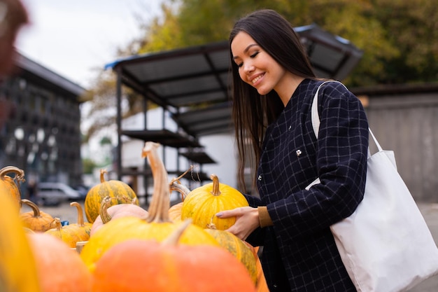 Giovane donna casual attraente che compra felicemente la zucca per il giorno di Halloween al negozio della fattoria di autunno all'aperto