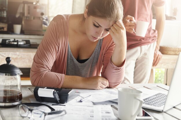 Giovane donna casual che ha uno sguardo depresso mentre gestisce le finanze della famiglia e fa le scartoffie, seduto al tavolo della cucina con un sacco di carte, calcolatrice e laptop