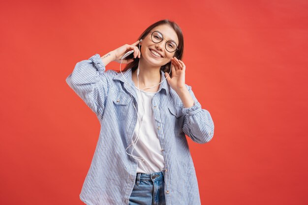 Giovane donna casuale che sorride con le cuffie e i vetri sulla parete rossa