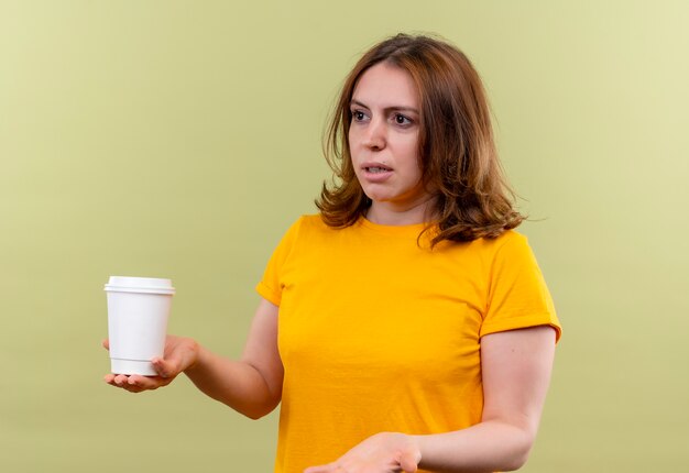 Giovane donna casuale dispiaciuta che tiene la tazza di caffè di plastica e che mostra la mano vuota sulla parete verde isolata