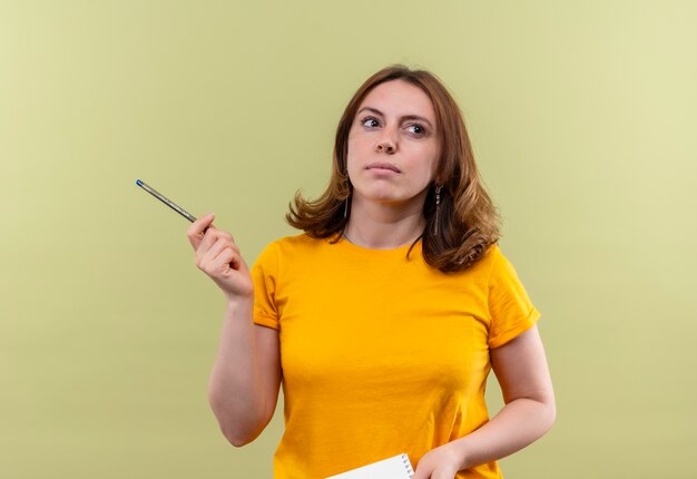 Giovane donna casuale premurosa che tiene la penna e il blocco note sulla parete verde isolata