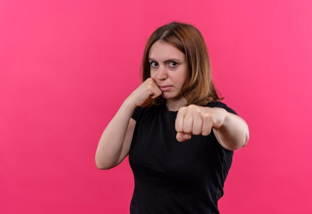 Giovane donna casuale sicura che allunga il pugno con e che mette un altro sulla guancia sullo spazio rosa isolato con lo spazio della copia