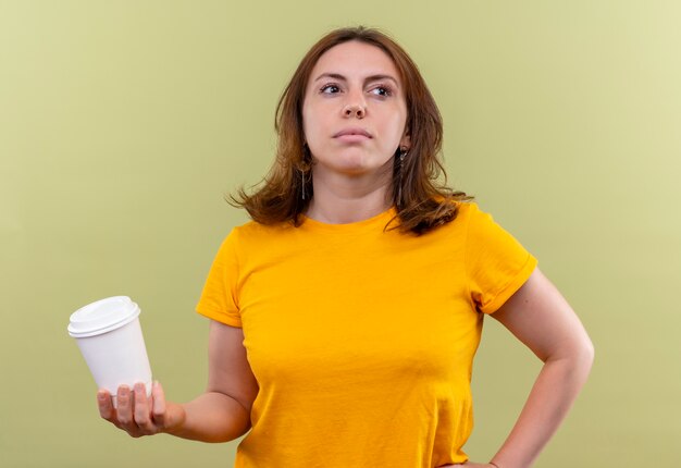 Giovane donna casuale sicura che tiene la tazza di caffè di plastica con la mano sulla vita su spazio verde isolato