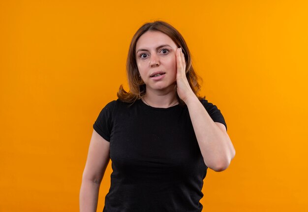 Giovane donna casuale sorpresa che mette la mano sulla guancia sulla parete arancione isolata con lo spazio della copia