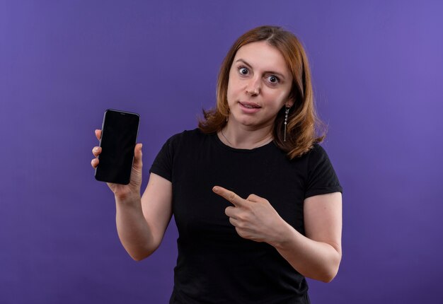 Giovane donna casuale sorpresa che tiene il telefono cellulare e indicandolo sulla parete viola isolata