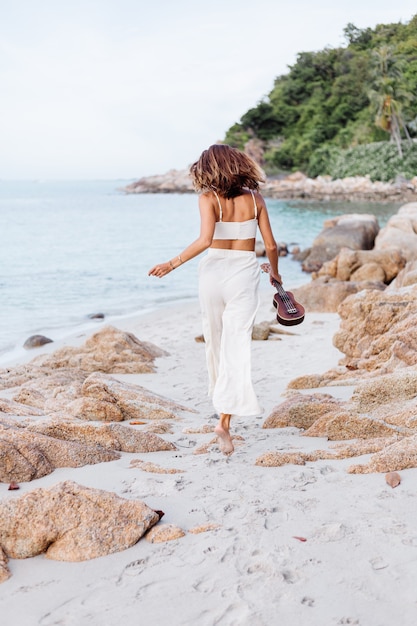 giovane donna caucasica calma felice con ukulele sulla spiaggia rocciosa tropicale al tramonto