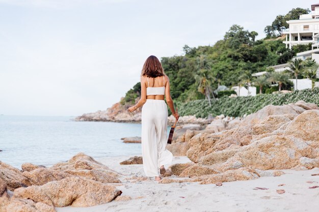 giovane donna caucasica calma felice con ukulele sulla spiaggia rocciosa tropicale al tramonto