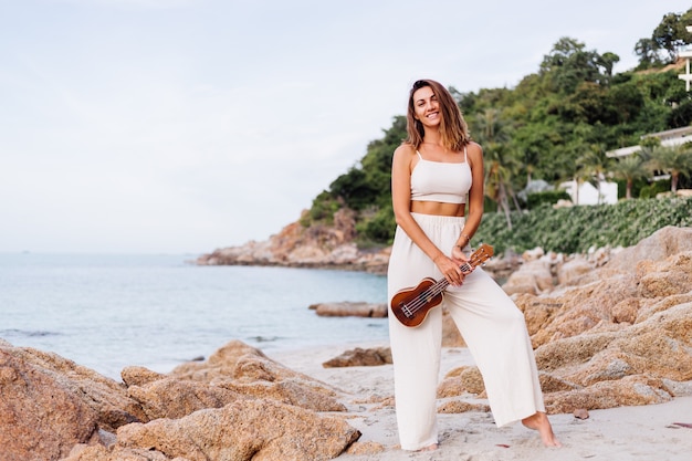 giovane donna caucasica calma felice con ukulele sulla spiaggia rocciosa tropicale al tramonto